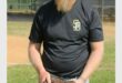 A white male with a long beard wearing a black shirt, white pants, a green cap and holding a baseball mitt.