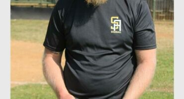 A white male with a long beard wearing a black shirt, white pants, a green cap and holding a baseball mitt.