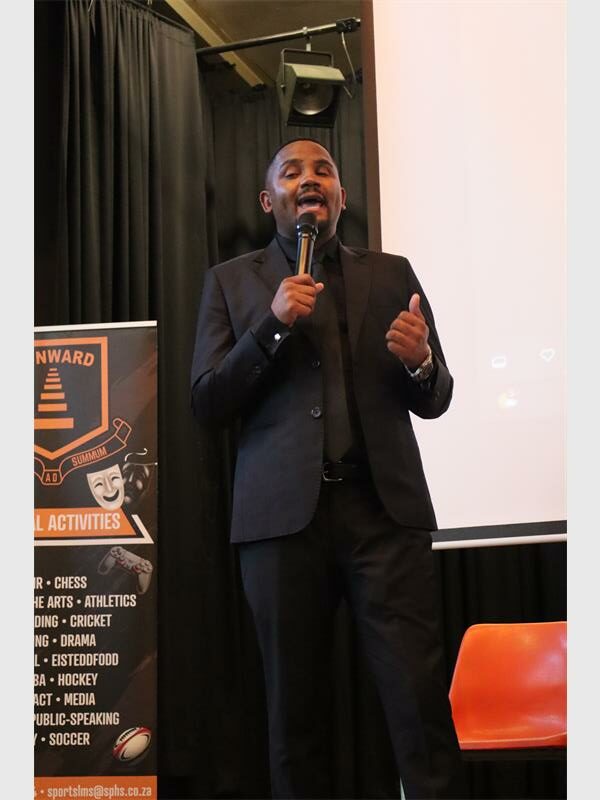 A black male on a school stage speaking into a microphone wearing a black suit, shirt and tie.
