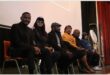 A group of female and male dignitaries sitting on chairs on the stage at a school.