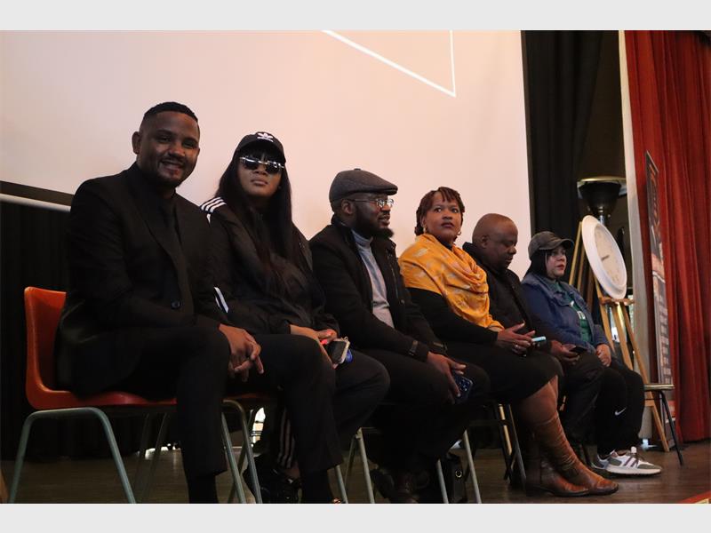 A group of female and male dignitaries sitting on chairs on the stage at a school.
