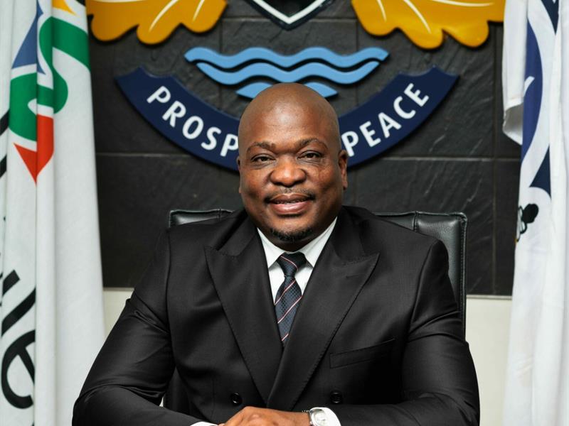A black man in a black jacket and tie sitting with his arms crossed at a desk with a logo behind him.