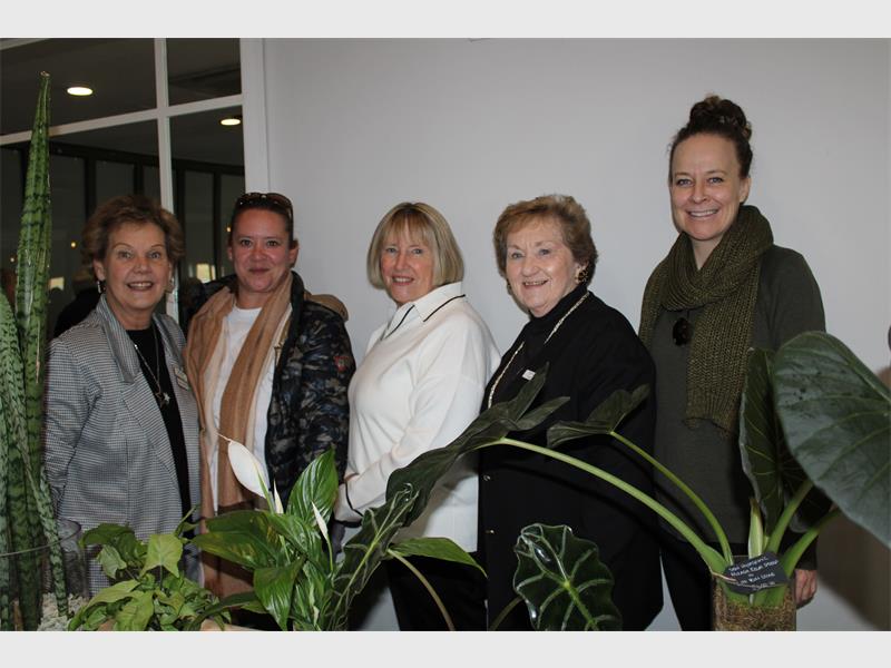 Five white women standing in a row in front of a white wall with leaves of green plants visible in front of them.