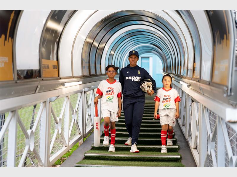 An adult cricket player flanked by two junior cricket players in KFC kit walking down steps through a tunnel to a cricket field.