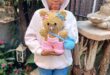 A young black girl wearing a white hoodie and jeans standing in a garden holding a handmade knitted teddy bear.