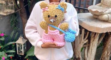 A young black girl wearing a white hoodie and jeans standing in a garden holding a handmade knitted teddy bear.
