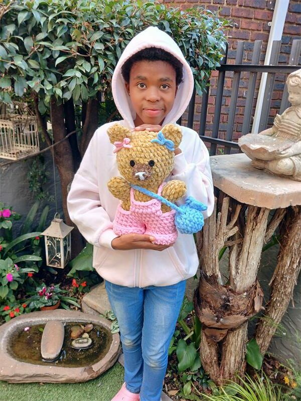 A young black girl wearing a white hoodie and jeans standing in a garden holding a handmade knitted teddy bear.