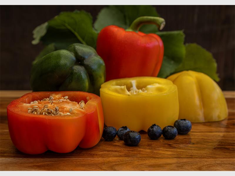 Yellow and red peppers displayed in cut format and whole on a wooden table with black berries in the foreground.