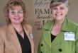 Two smiling women stand together, one in a tan blazer and glasses, the other in a green coat. They are in front of a sign with inspirational text.