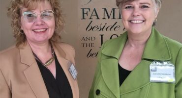 Two smiling women stand together, one in a tan blazer and glasses, the other in a green coat. They are in front of a sign with inspirational text.
