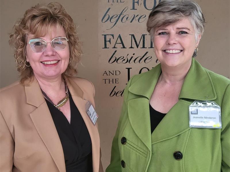 Two smiling women stand together, one in a tan blazer and glasses, the other in a green coat. They are in front of a sign with inspirational text.