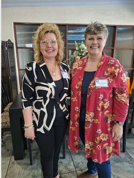 Two women, one dressed in black and white and the other in a black top with a read floral shirt over and both wearing name badges.
