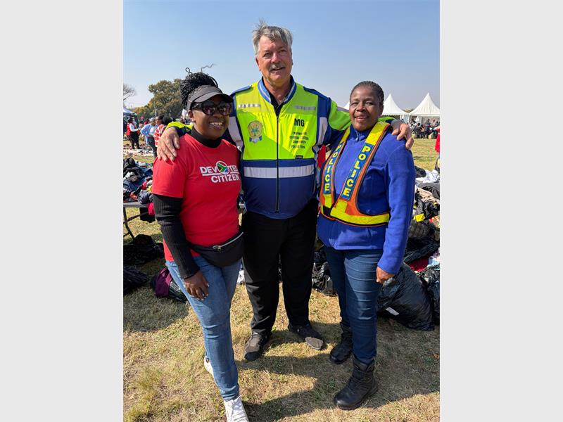 A white male wearing a reflective vest flanked by two black females, one in red shirt and one in a SAPS reflective.