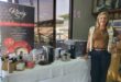 A blonde woman dressed in jeans and a tan top standing next to a table displaying coffee machines and accessories.