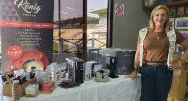 A blonde woman dressed in jeans and a tan top standing next to a table displaying coffee machines and accessories.