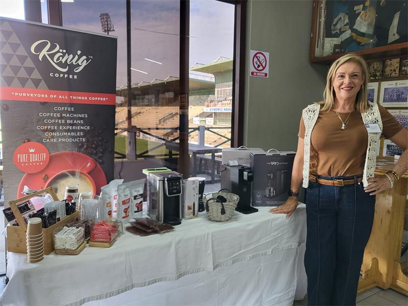 A blonde woman dressed in jeans and a tan top standing next to a table displaying coffee machines and accessories.