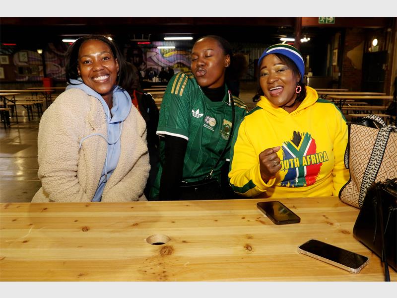 Three black females sitting at a wooden table, two dressed in Bafana Bafana shirts and the other in a light coloured jacket, smiling for the camera.