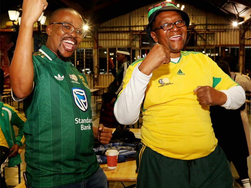 Two black males dressed in Bafana Bafana shirts cheering with their hands raised.
