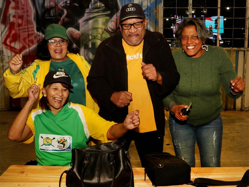 Three black women and a black man cheering in Bafana Bafana shirts.