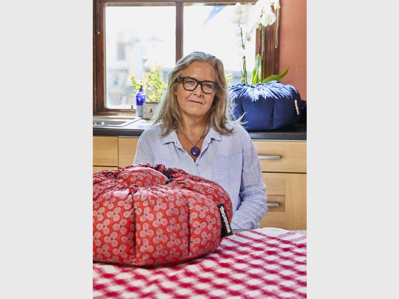 A white woman with long blonde hair and glasses sitting at a table with a checkered red and white table cloth on it and a red African print Wonderbag.