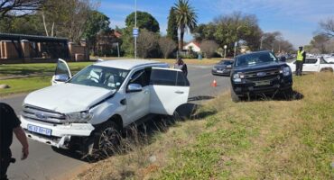 A crime scene showing a white bakkie and a darker bakkie in the background parked on the side of a road with the white bakkie's back doors open.