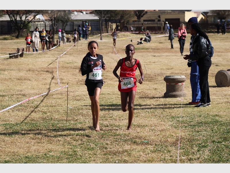 Two young black athletes running on a grass track towards a finish line.