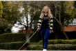 A young white girl with long blonde hair wearing a striped open jersey, brown shirt and blue jeans and walking with a white cane.