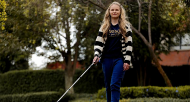 A young white girl with long blonde hair wearing a striped open jersey, brown shirt and blue jeans and walking with a white cane.