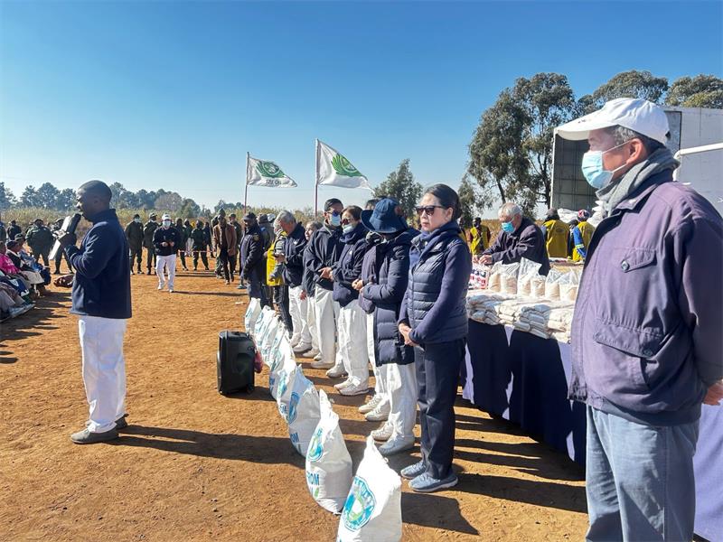 Members of a NPO all dressed in white pants and blue jackets standing in row with food parcels at their feet with one of the members in front of them addressing a crowd on a microphone.