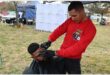 Two males, one sitting on a chair with a black hairdressing cape on having his hair shaved by a man wearing a red shirt.