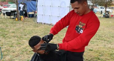 Two males, one sitting on a chair with a black hairdressing cape on having his hair shaved by a man wearing a red shirt.