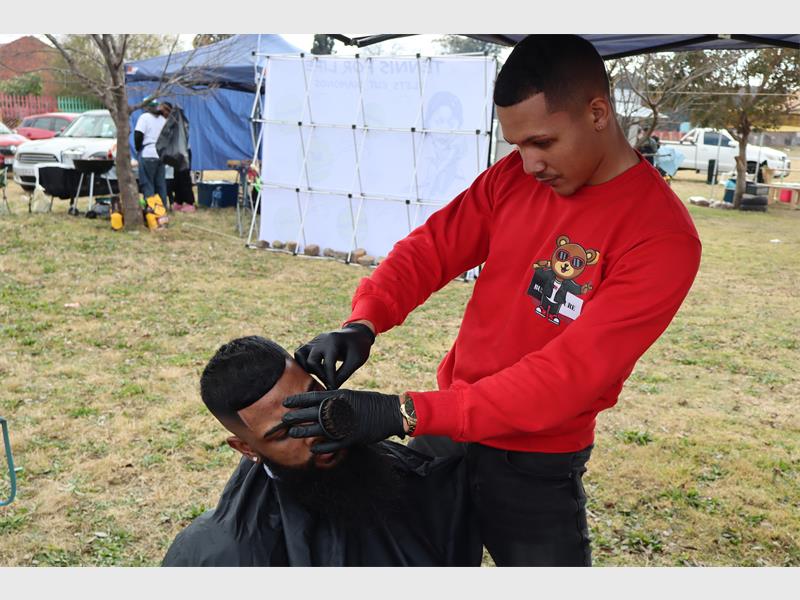 Two males, one sitting on a chair with a black hairdressing cape on having his hair shaved by a man wearing a red shirt.