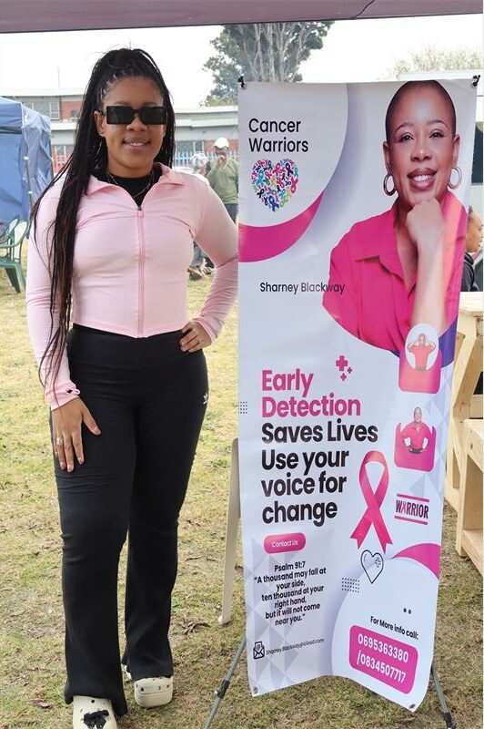 Lady in a pink top and black tights standing next to a banner with pink cancer ribbons and information on it.