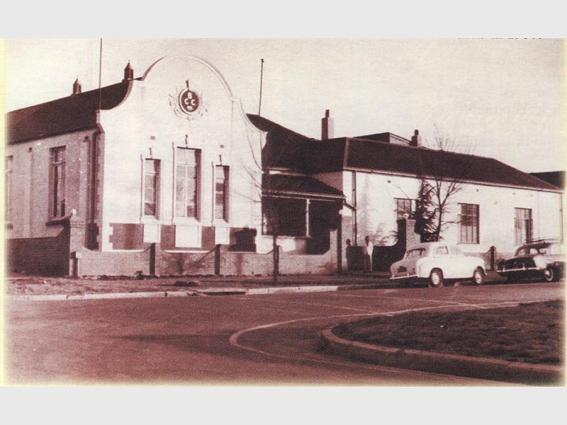 A black and white image of an old building with an old white car parked outside on the street.
