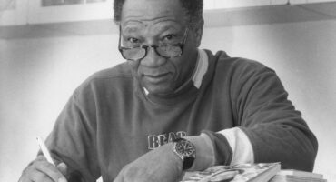 A black man looking over a pair of reading glasses, with his arm resting on a table and a book.