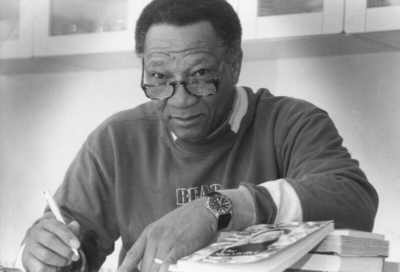 A black man looking over a pair of reading glasses, with his arm resting on a table and a book.