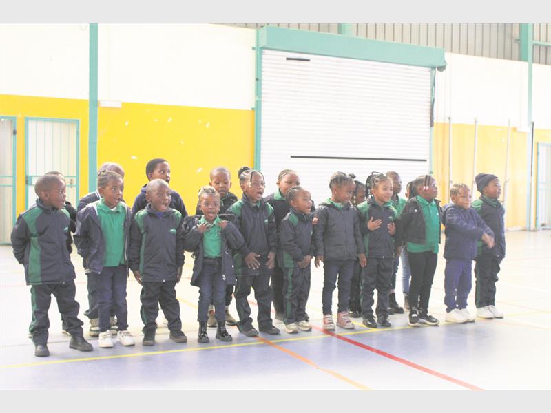 A group of small children dressed in black tracksuits and green shirts standing in a row in a hall.