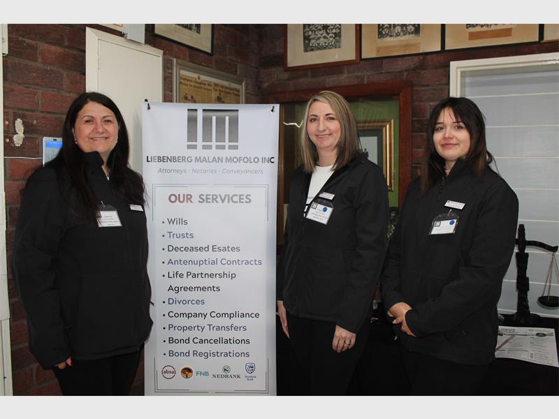 Three women standing either side of a banner, all wearing black suits.