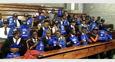 A group of male learners sitting in a hall wearing blue school uniform and holding bags containing promotional items.