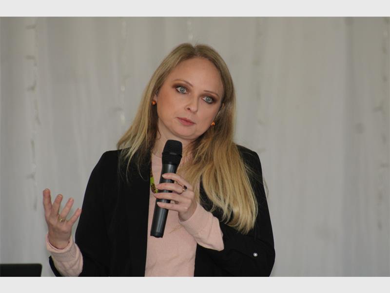A white woman with long blonde hair wearing a black jacket and speaking into a microphone.