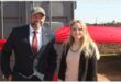 A white man in a suit next to a white woman with long blonde hair in front of a container with a big red ribbon around it.