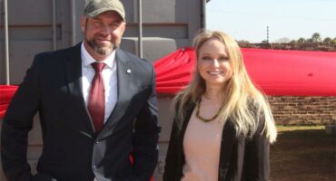 A white man in a suit next to a white woman with long blonde hair in front of a container with a big red ribbon around it.