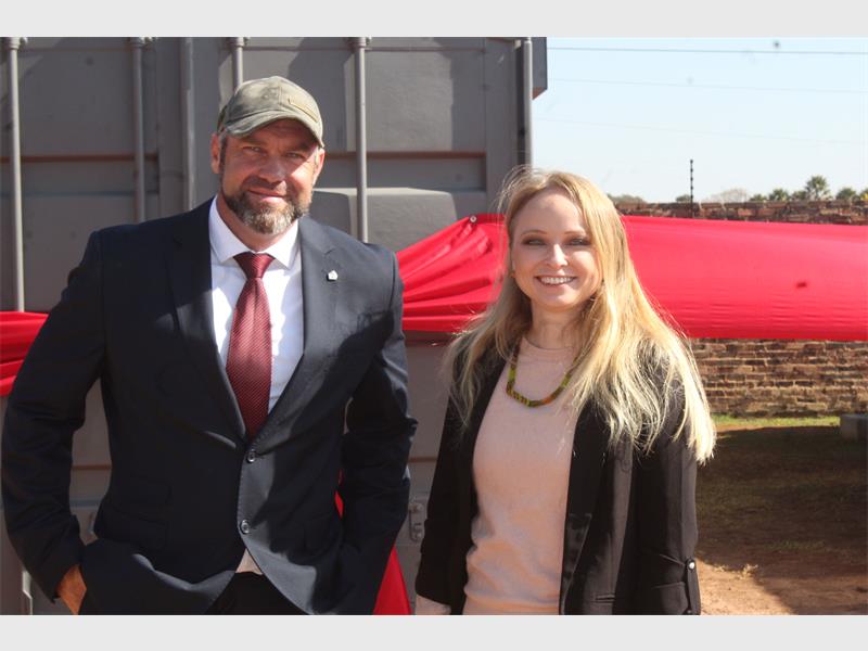 A white man in a suit next to a white woman with long blonde hair in front of a container with a big red ribbon around it.