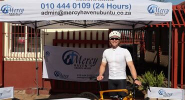 A white male standing behind a bicycle, dressed in cycling gear under an gazebo and next to a banner.