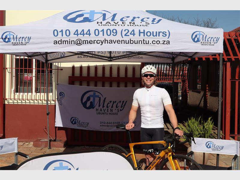 A white male standing behind a bicycle, dressed in cycling gear under an gazebo and next to a banner.