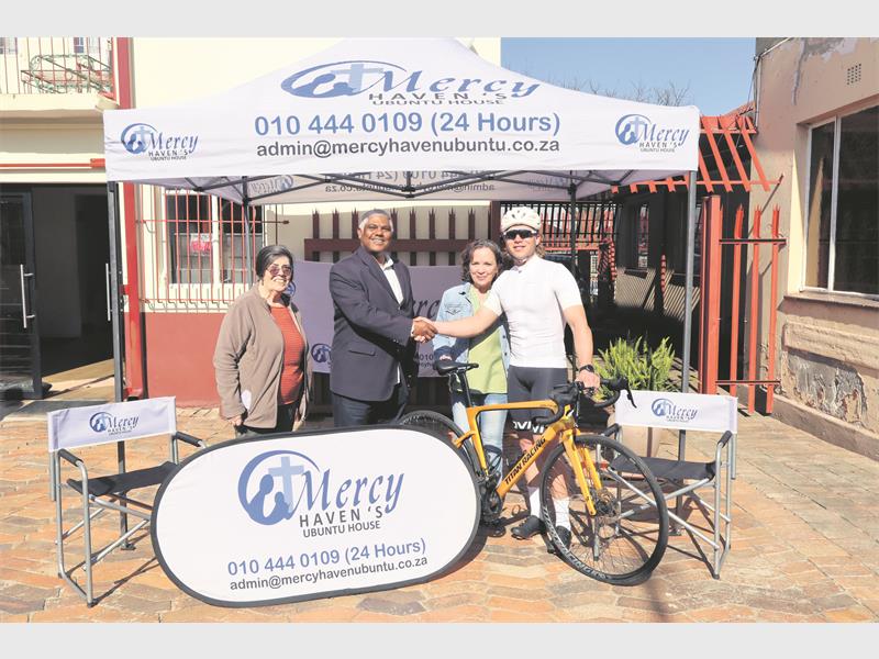 Two women and two men standing behind a white banner reading Mercy Haven and under a white gazebo with the same wording, the one man cycling gear and standing with a bicycle.