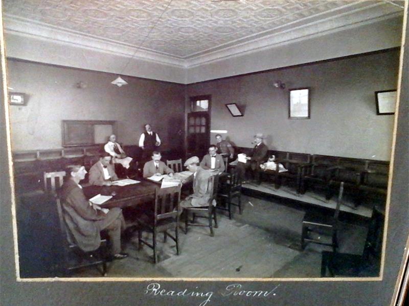 A black and white image of a reading room inside a community club with several men sitting at wooden tables.