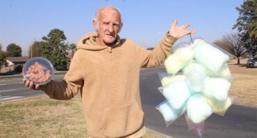 Elderly man standing on a street corner holding candyfloss and koeksisters