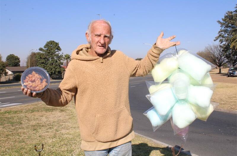 Elderly man standing on a street corner holding candyfloss and koeksisters