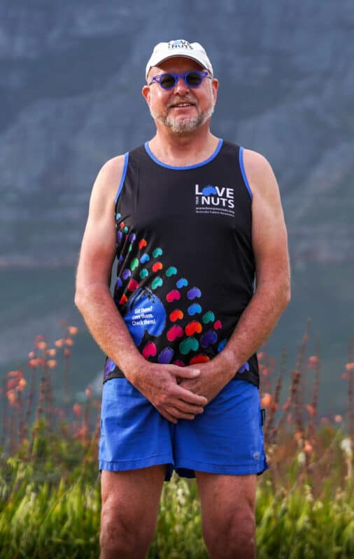 A white man dressed in blue shorts, a black shirt and a cap and wearing sunglasses with his hands clasped in front of his groin.